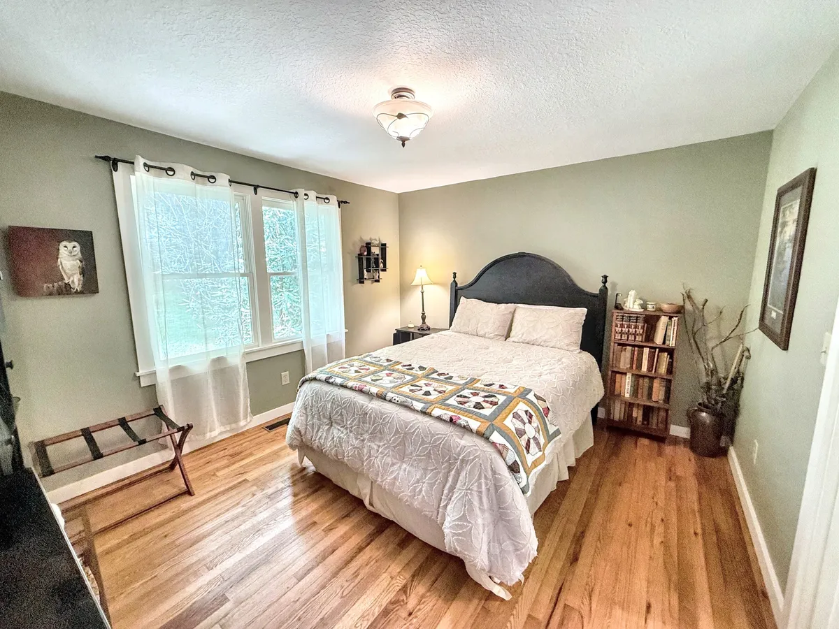 A sunlit bedroom nook with a bed topped with a quilt, a nearby bookshelf, a lamp, and large windows that let in natural light. The space exudes warmth and invites moments of relaxation or quiet conversation.