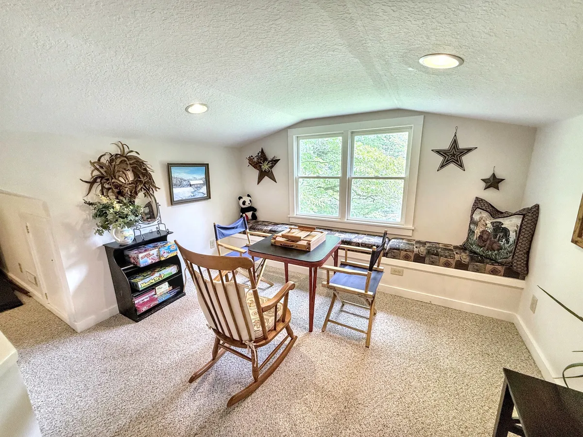 A tranquil nook featuring a table stacked with books, a rocking chair, a window seat, and decorative wall art. Sunlight streams through the window, creating an inspiring spot for reading, games, or creative pursuits.