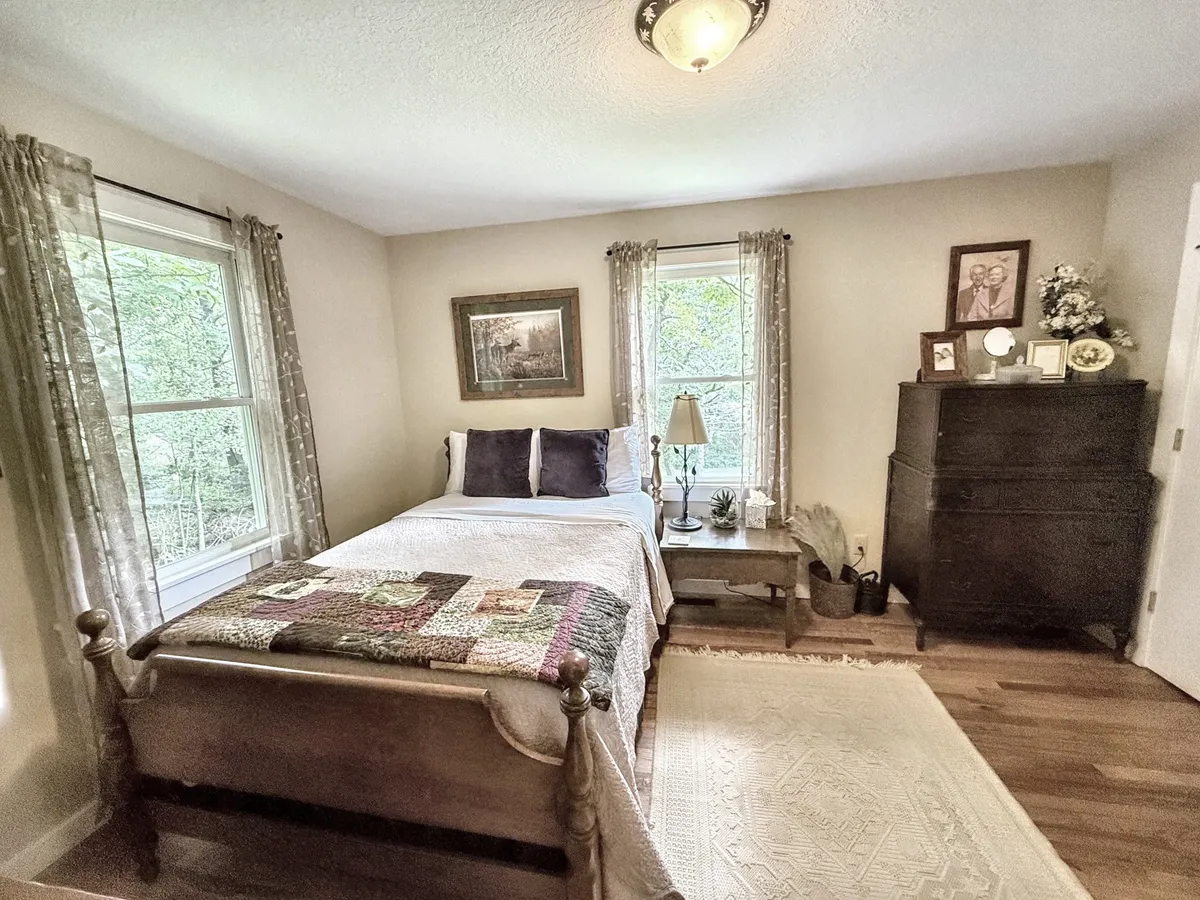 A bright queen bedroom with a plush bed, a sunlit window, a dresser, and thoughtfully arranged artwork. The space invites relaxation and quiet moments, perfect for restful nights and peaceful mornings.