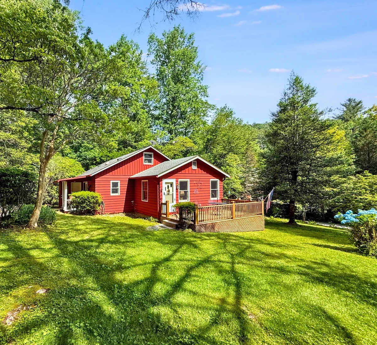 The image features a storybook-red cottage nestled amid mature trees, surrounded by a lush green lawn. A spacious wooden deck invites guests to relax, with sweeping views of the peaceful natural landscape. The setting glows in soft, welcoming light—ideal for sunset gatherings or morning coffee outdoors.