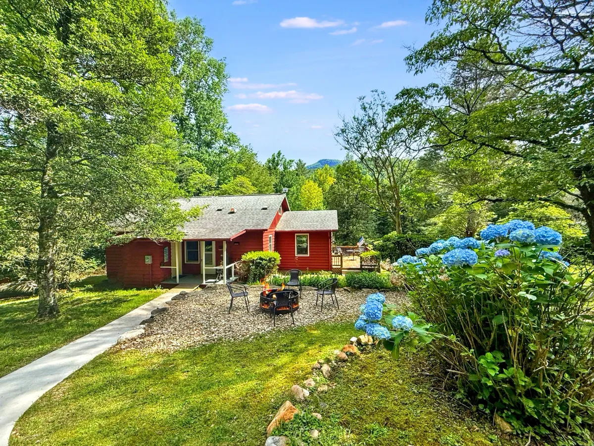 This exterior image features the red house surrounded by lush greenery and blooming blue hydrangeas. The inviting fire pit area with outdoor seating is visible in the foreground, with hints of a serene mountain backdrop. The scene is bathed in soft evening light, suggesting opportunities for relaxing outside under the stars.