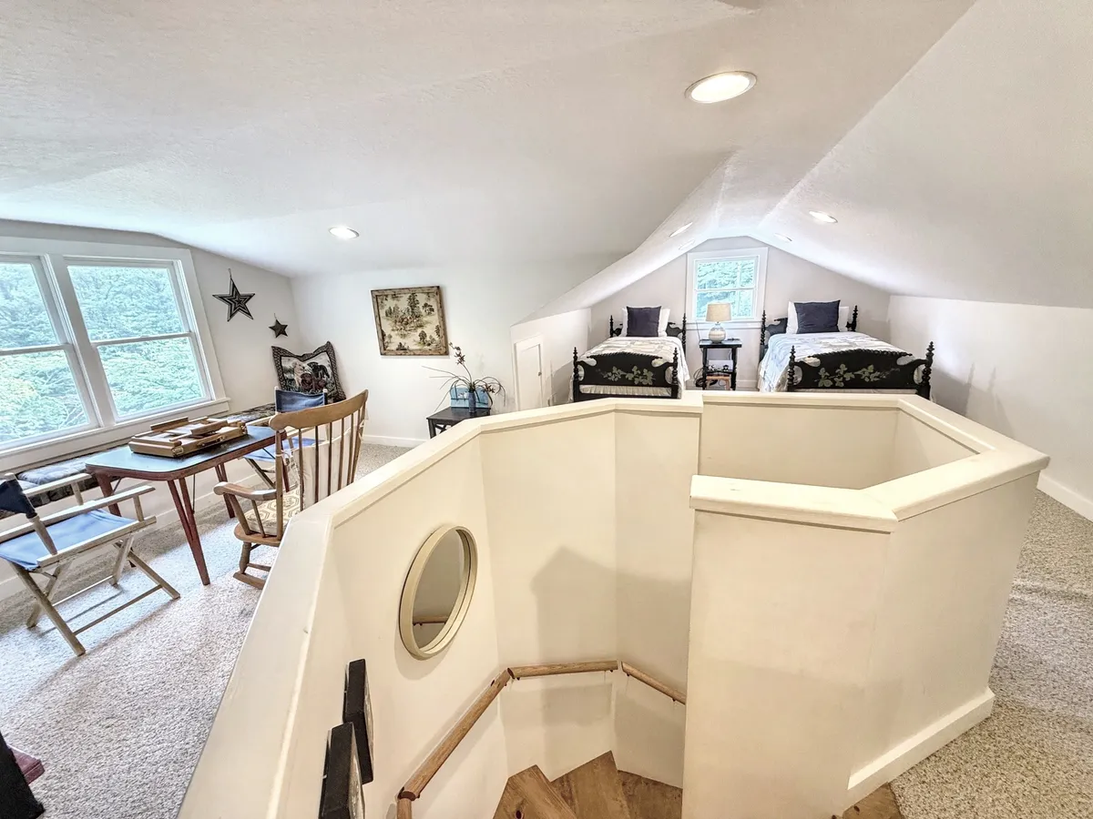 This image features a bright loft space with two beds dressed in floral bedding, a wooden table with chairs set for crafts or games, and a stylish staircase with a circular design. A decorative mirror adds character, and the cozy setting is flooded with natural light, perfect for both children and adults.
