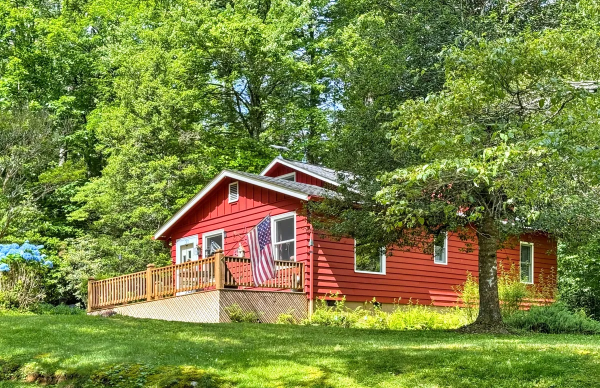 The image shows the red house from another exterior angle, highlighting its welcoming deck draped in lush greenery and seasonal flowers. An American flag waves gently on the porch, and the deck is set up for relaxing and enjoying the outdoors. The natural landscape frames the house, reinforcing its tranquil setting.