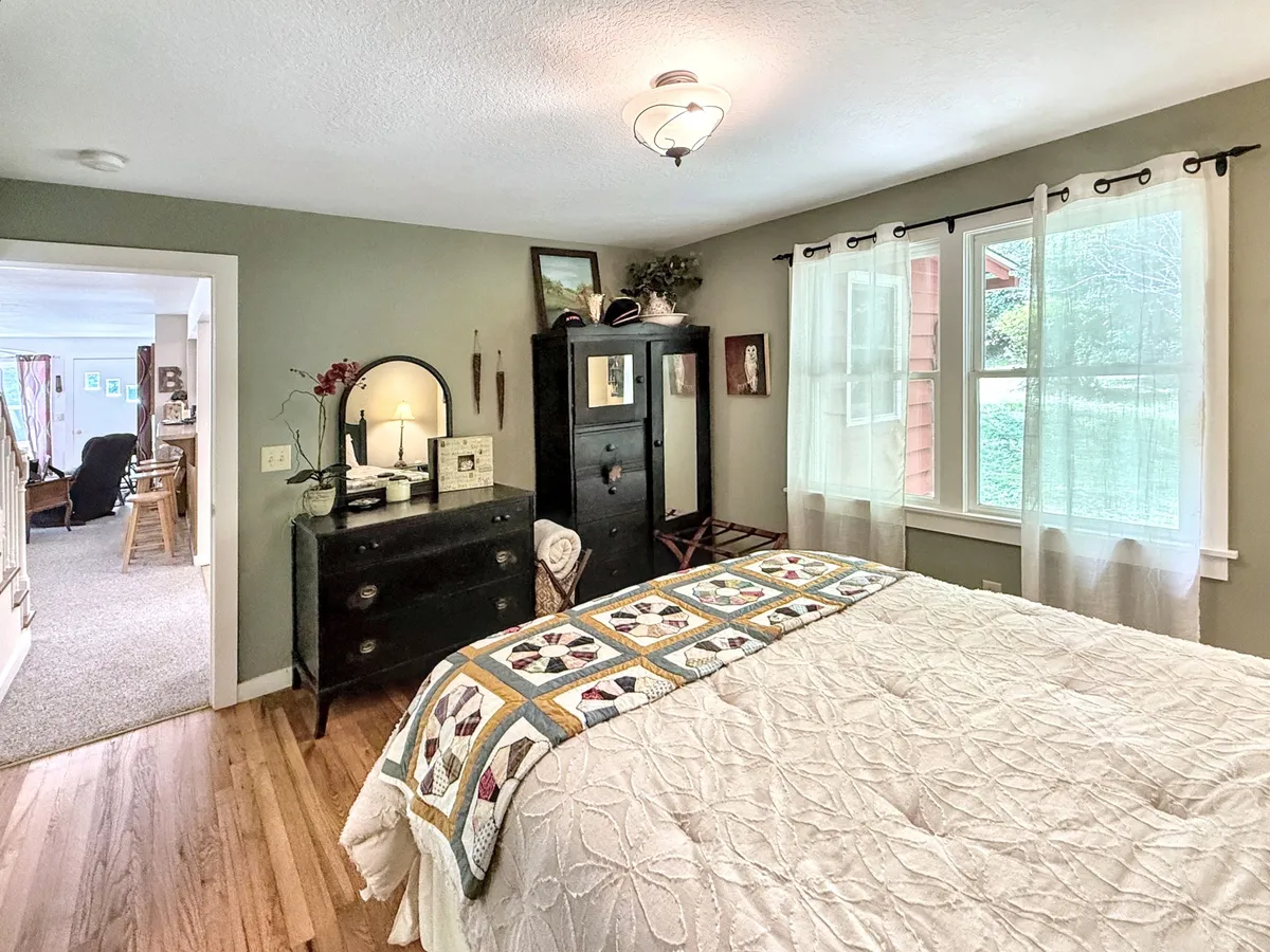 A cozy bedroom featuring a bed adorned with a decorative quilt, a window draped with sheer curtains allowing gentle natural light, and a dresser with a mirror. The room exudes warmth and comfort, ideal for reading or unwinding.