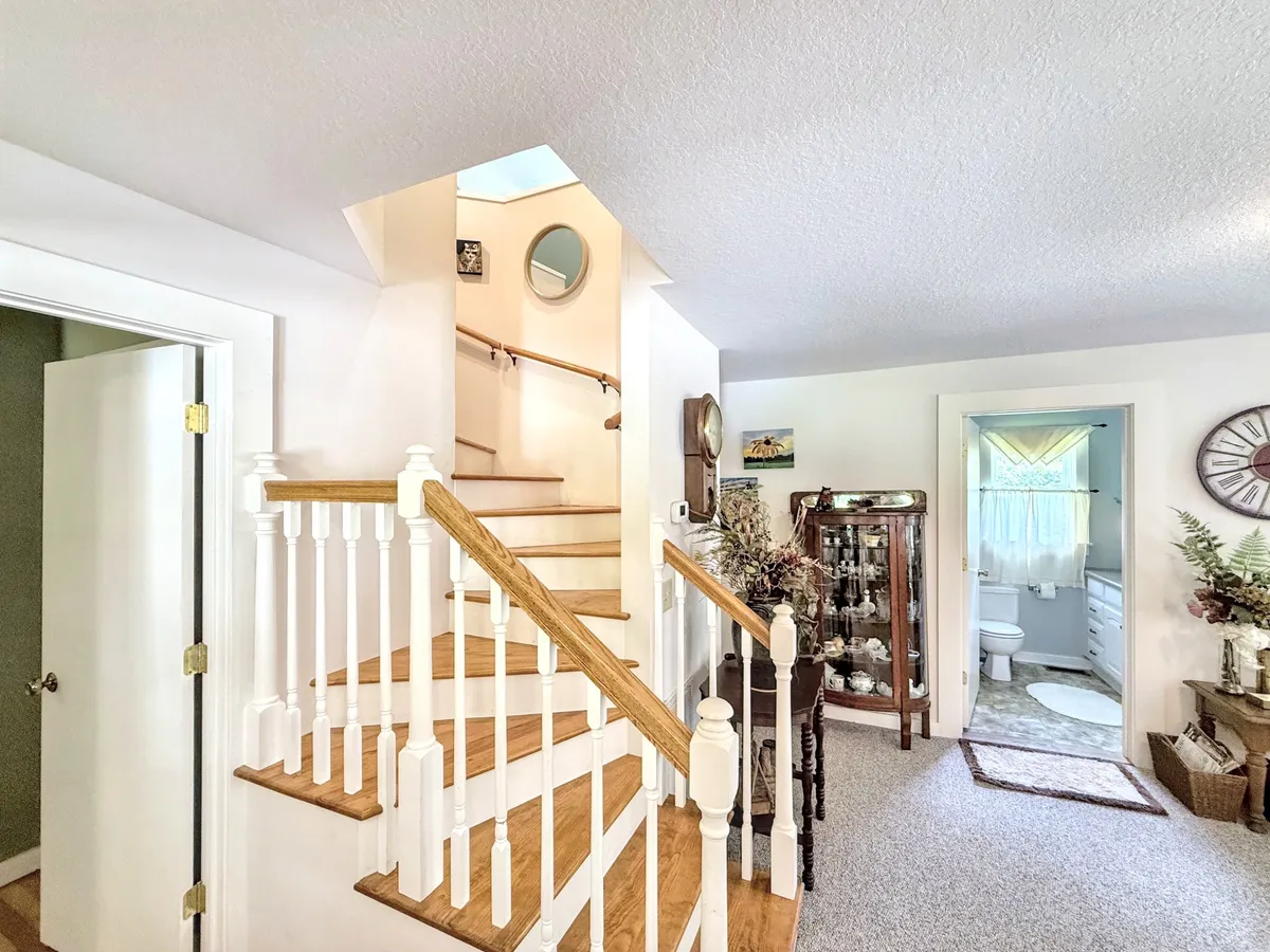 A bright hallway featuring a staircase leading to the upper loft, a decorative cabinet that hints at family history, and a visible bathroom door. Light pours in through a skylight, enhancing the sense of welcome and openness.